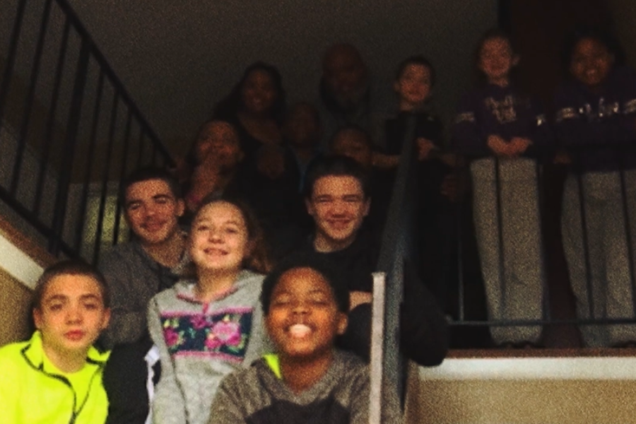 A large group of children and adults pose together on a staircase inside a dimly lit home.
