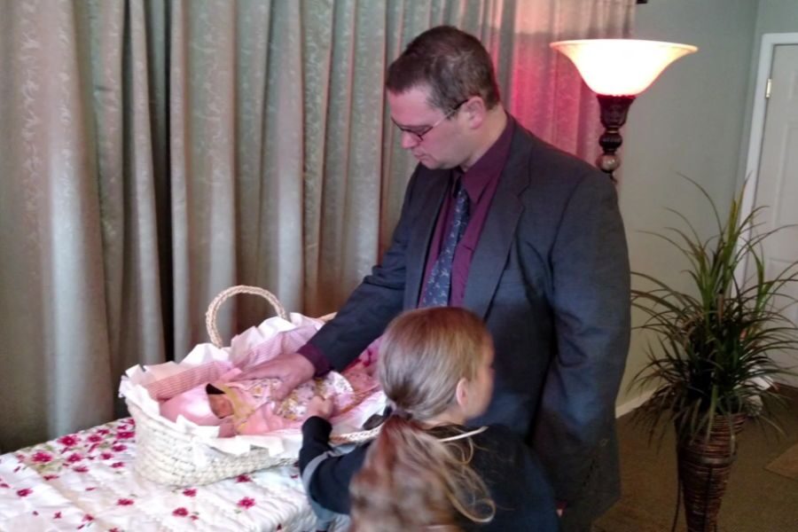 A man in a suit gently touches a baby in a white wicker casket lined with pink fabric. A young girl stands beside him looking at the baby.