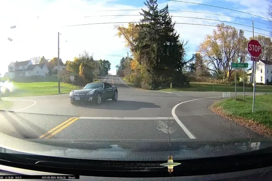 Dashcam view of a black convertible turning left in front of an oncoming car at a rural intersection with stop signs.