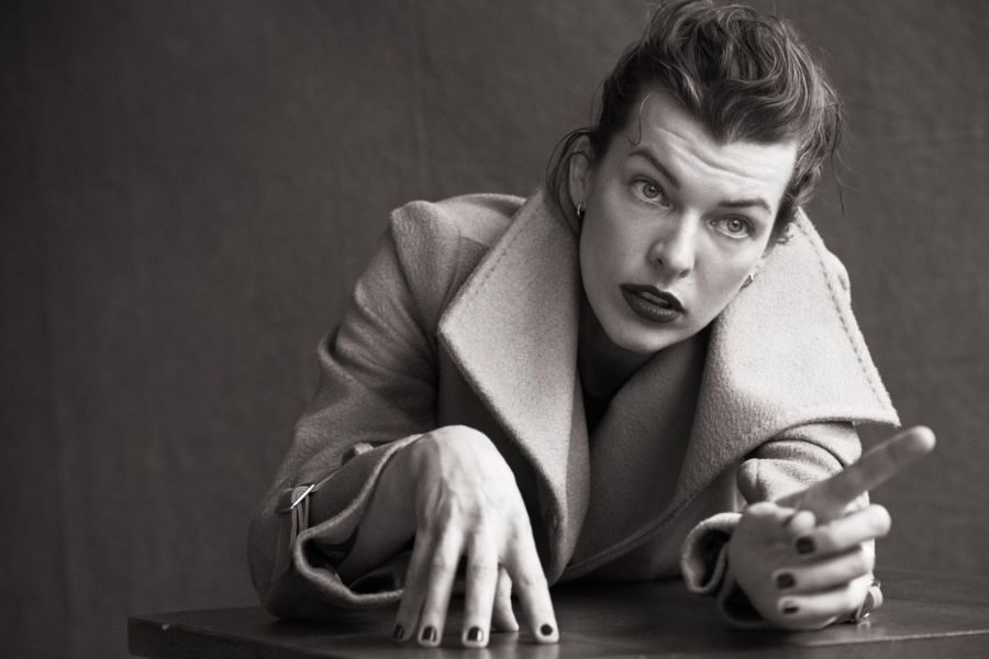 A black-and-white portrait of a model in a wool coat leaning forward with a strong gaze and expressive hands.