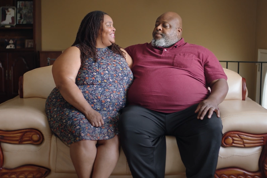 A couple sitting close together on a tan couch in their home, looking at each other and smiling.