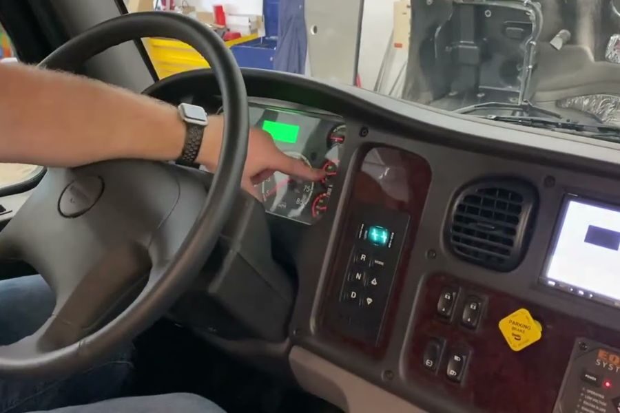 Close-up of a man’s arm inside a large vehicle, pointing at gauges on the dashboard while seated at the steering wheel.
