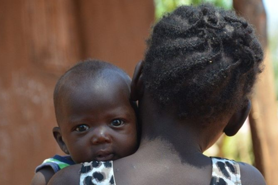 A baby with wide eyes looks over the shoulder of an older girl with braided hair, being held closely.