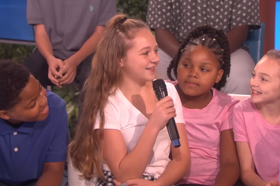 A girl holding a microphone smiles as she speaks on a talk show, surrounded by other smiling children.