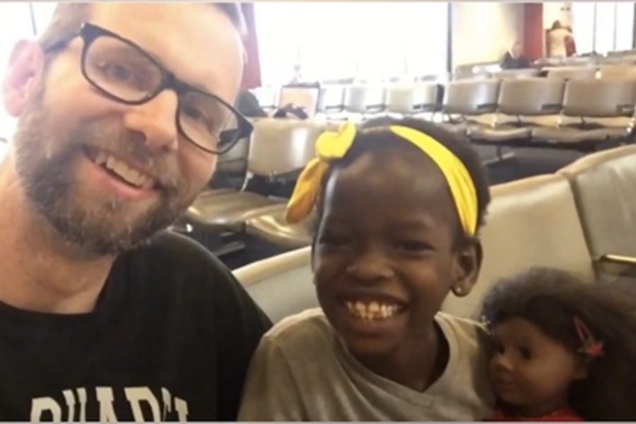 A man and young girl with a yellow headband smile for a selfie in an airport waiting area with empty chairs.