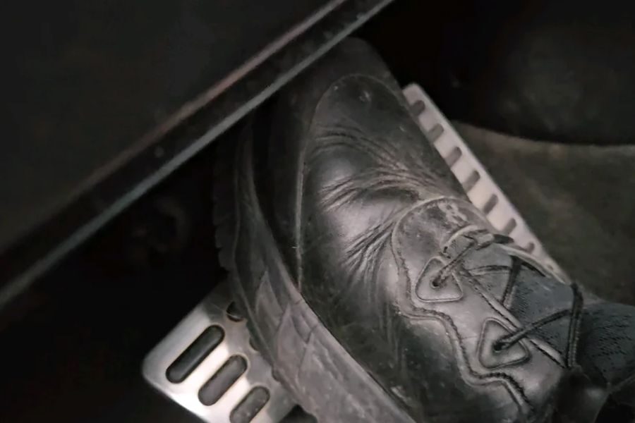 Close-up of a black shoe pressing down on a car’s gas pedal with a metallic footrest beside it.