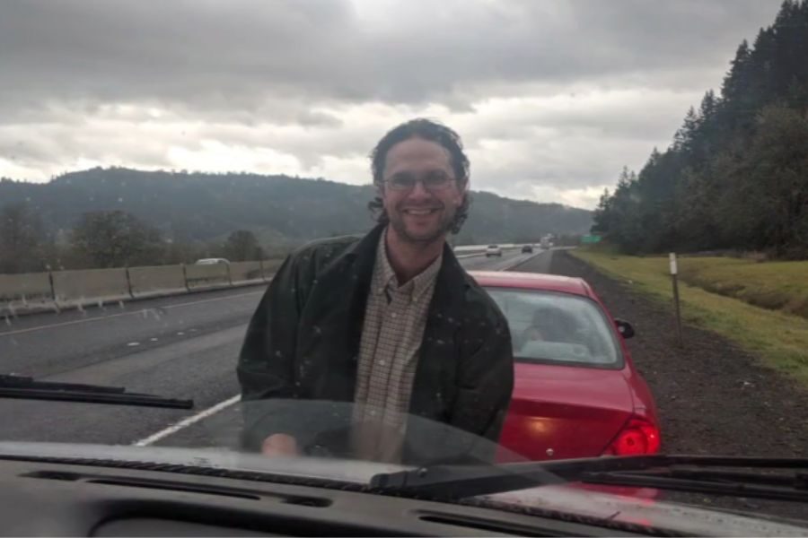 A man smiles while standing in front of a red car on the shoulder of a highway with overcast skies and forested hills in the background.