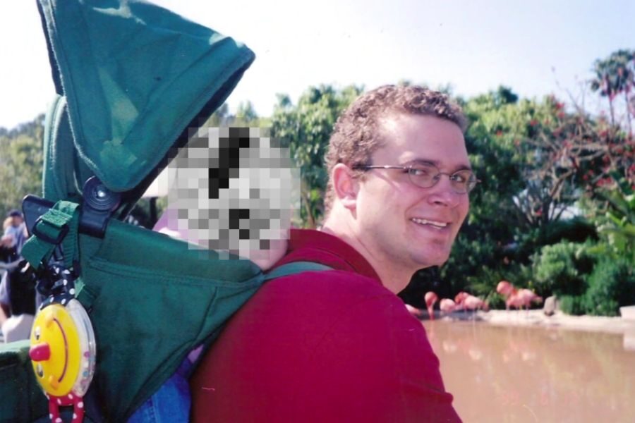 A man wearing glasses and a red shirt carries a young child in a green backpack carrier at a zoo, with flamingos and trees visible in the background. The child's face is pixelated.