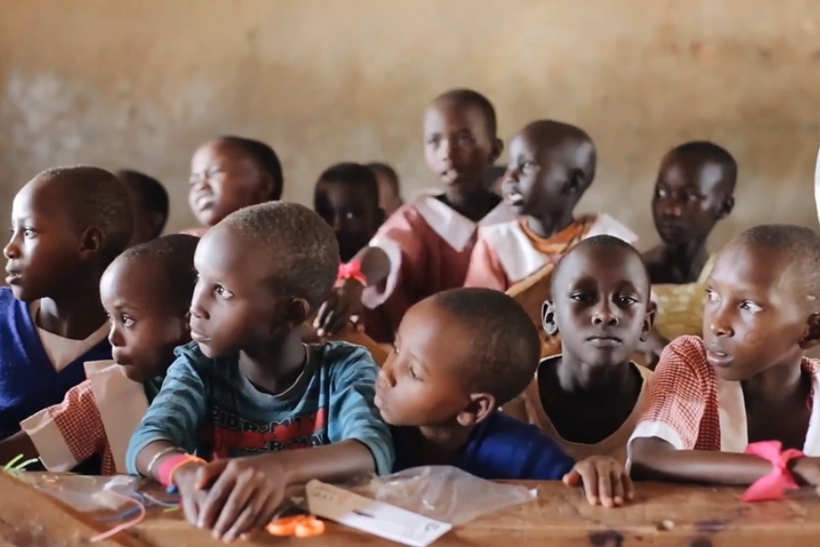 A crowded classroom with young children seated at wooden desks, many looking to the side in curiosity or distraction.