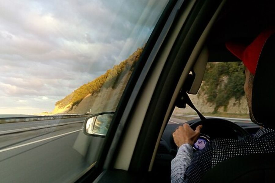 View from inside a car showing a man driving along a coastal highway with cliffs and the sea to the left.
