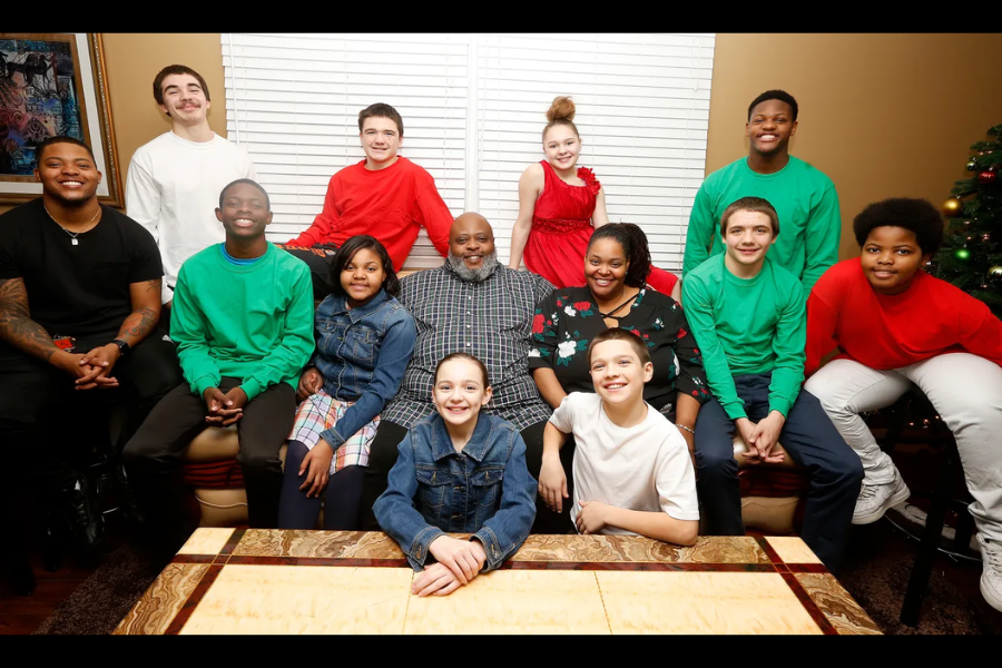 A large family dressed in festive red, green, and denim outfits posing together in a warmly lit living room for a holiday photo.