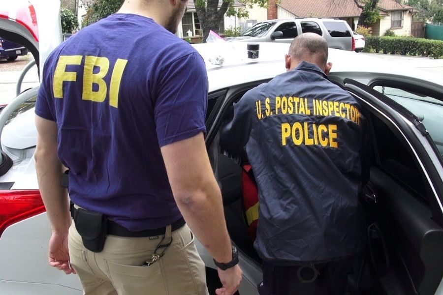 Two law enforcement officers, one in an FBI shirt and the other labeled U.S. Postal Inspector Police, stand by an open car door during an investigation.