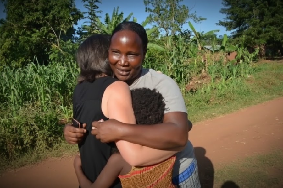 A woman warmly hugs another woman and child on a rural dirt path surrounded by lush greenery.