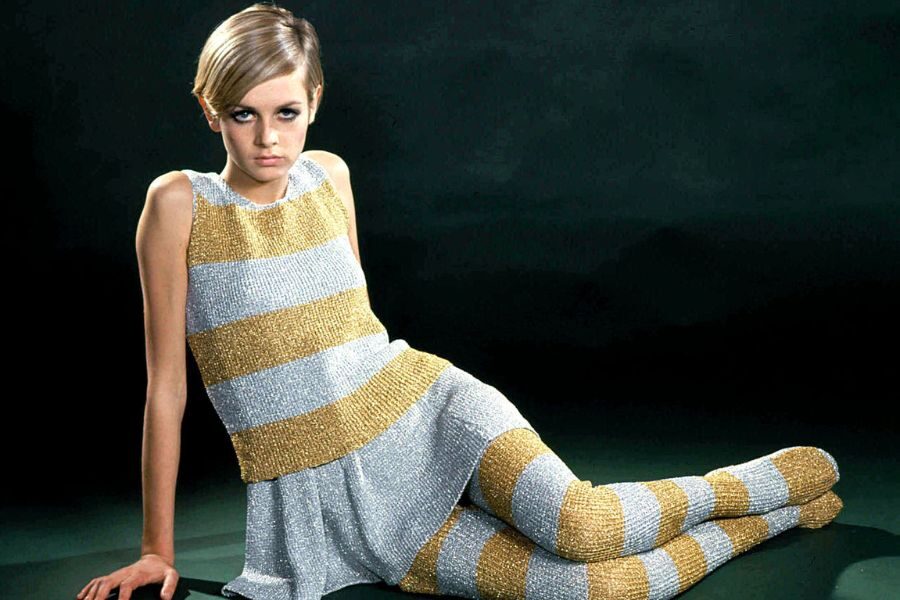 A model with a pixie haircut wears a glittering silver and gold striped outfit with matching leggings while seated dramatically.
