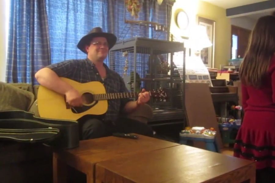 A man in a hat plays an acoustic guitar in a cozy living room while a child in a red dress stands nearby.