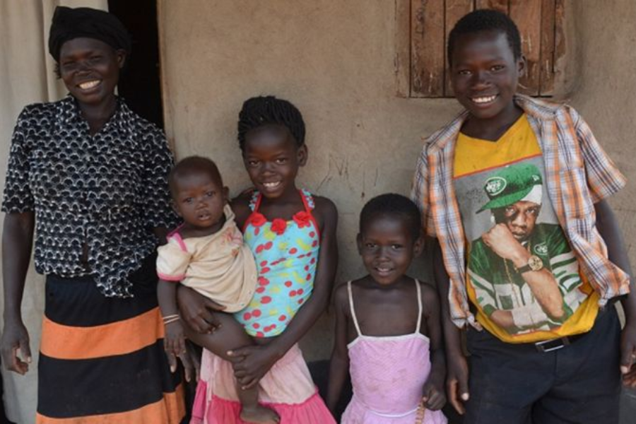 A smiling woman stands with four children outside a mud-brick home, one child is holding a baby.