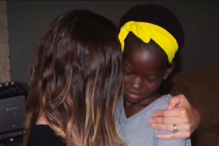 A young girl with a yellow headband receives a comforting hug from a woman with long brown hair indoors.