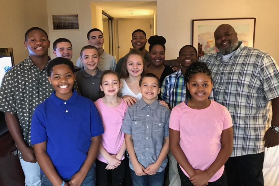 A diverse family of fifteen smiling together for a group photo in a hotel room with neutral decor.