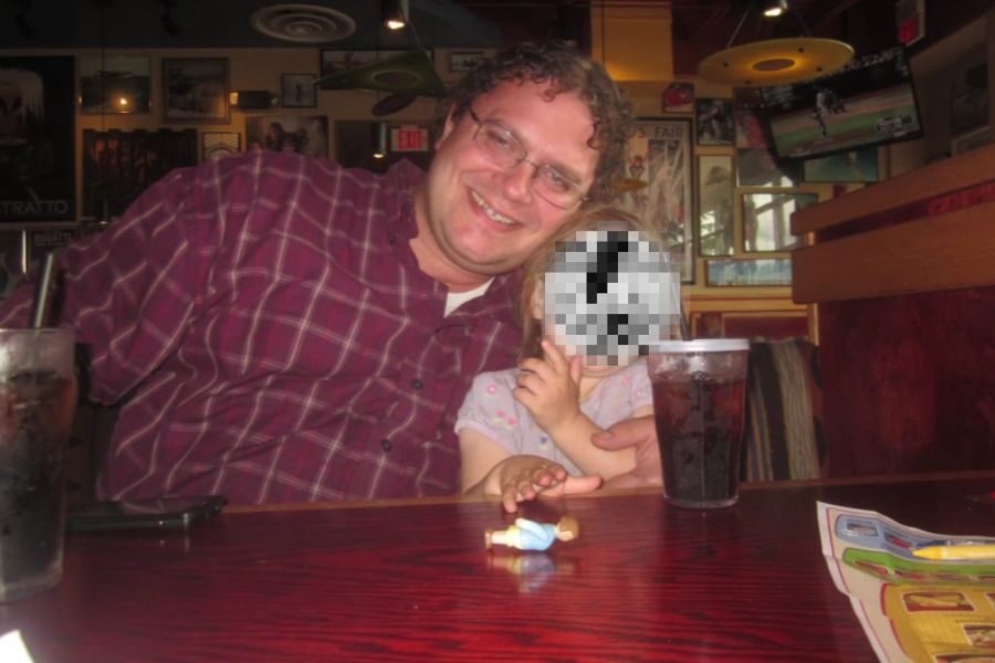 A smiling man in glasses hugs a young child with a pixelated face at a restaurant booth.