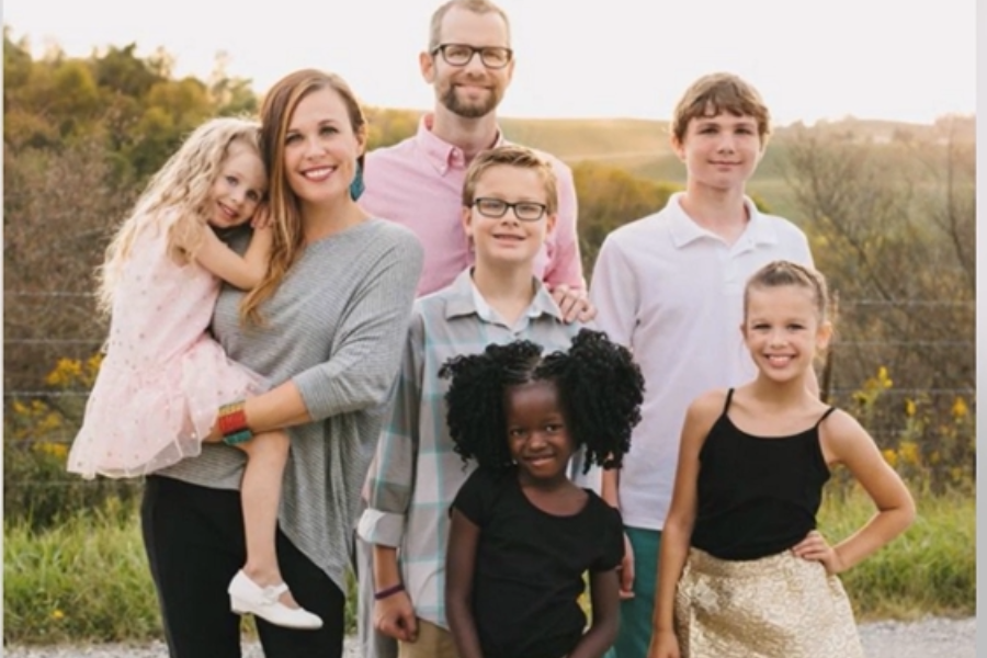 A family of seven poses for a portrait outdoors with smiles, including parents and children of different backgrounds.