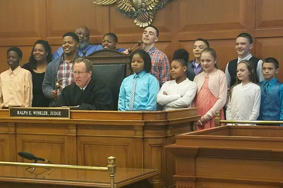 Parents of Five Decide to Bring Home Six More — And Let the ‘Chaos of Love’ Begin A group of children and adults gathered in a courtroom behind a judge named Ralph E. Winkler, who is seated at the bench with a gavel. Everyone is smiling and dressed formally.