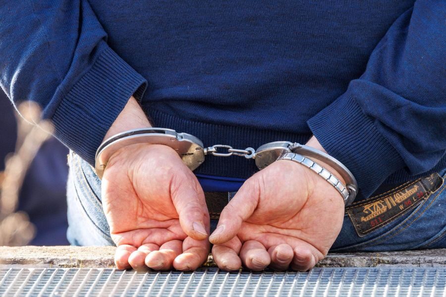 A close-up of a man’s hands behind his back, secured in handcuffs. He is wearing a blue sweater and jeans.