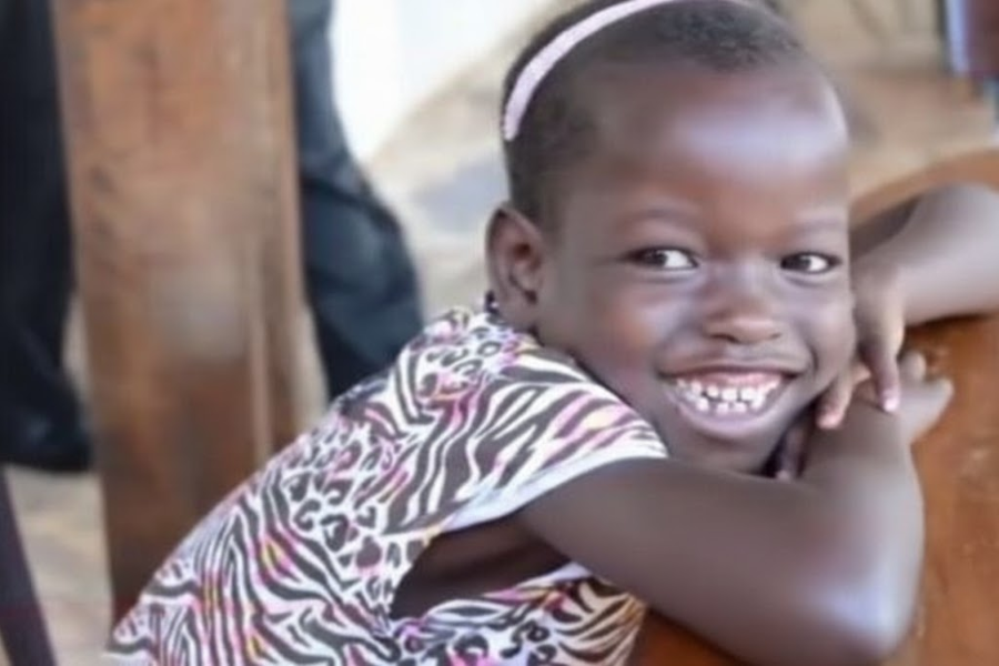 Family Adopts a 5-Year-Old — Then a Shocking Detail About Her Past Comes to Light A smiling young girl with a pink headband leans her arms on a table while wearing a zebra-print shirt.