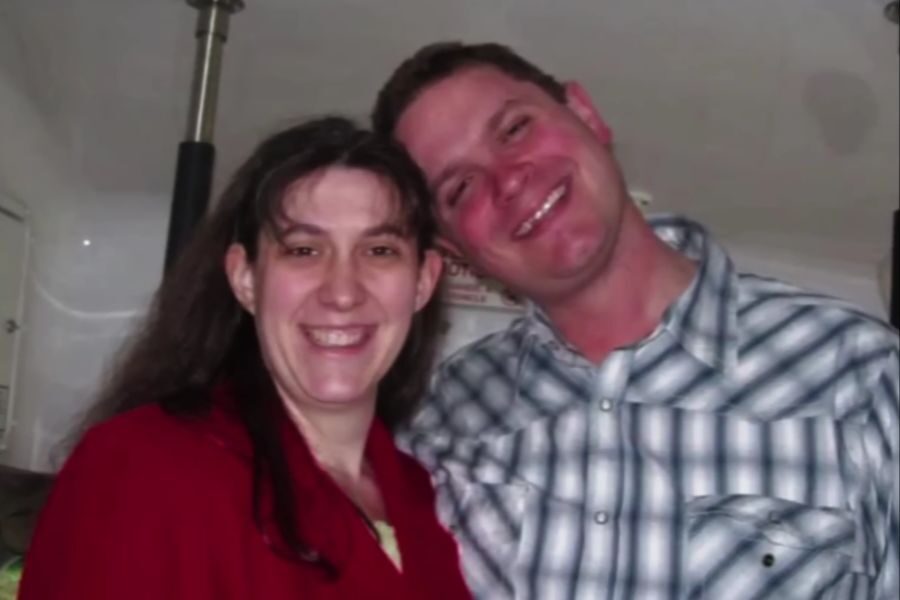 A couple smiles while standing closely together indoors. The man rests his head on the woman’s shoulder.
