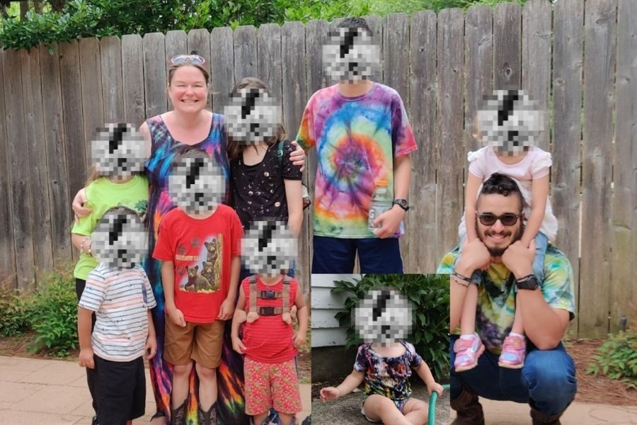 A woman and a man, both in tie-dye shirts, pose with a large group of children in front of a wooden fence. All children’s faces are pixelated.