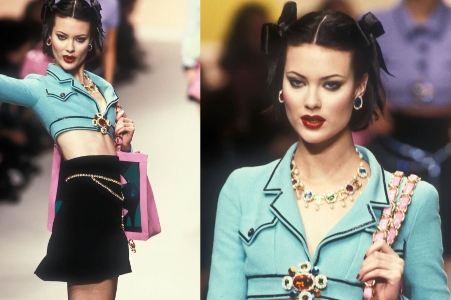 A model struts in a cropped turquoise jacket and black mini skirt, accessorized with a gem-studded belt and matching jewelry.