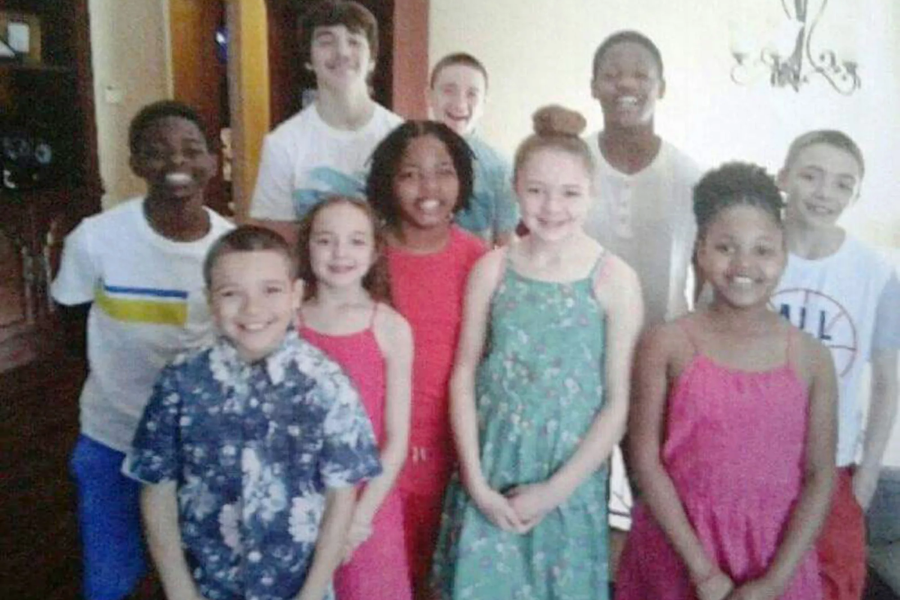 A group of twelve smiling children posing in a home setting, dressed in bright, colorful clothes.