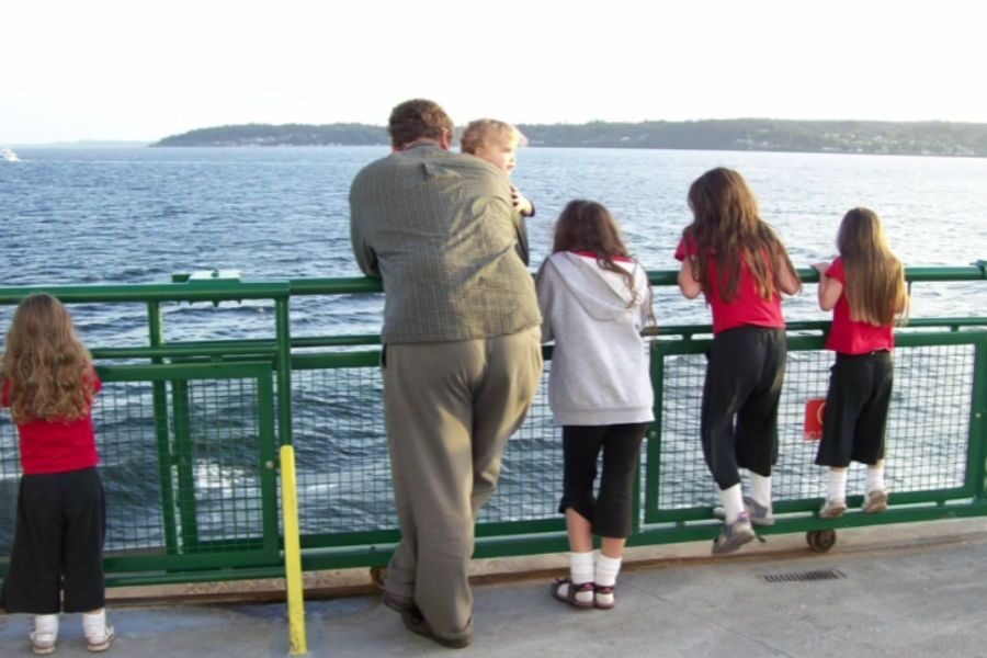 A man stands on a ferry deck overlooking the water with five children, some in matching red tops and black pants, leaning on the green railing.