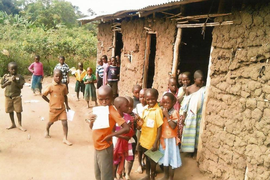 A large group of barefoot children holding papers and standing outside a mud-brick school building.