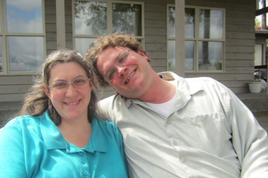 A smiling couple sits closely together on a bench outside a building with large windows.
