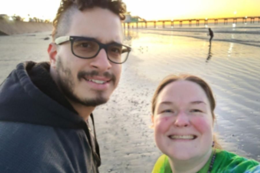 A smiling couple takes a selfie on a beach during sunset, with soft golden light reflecting off the water and a pier visible in the background.