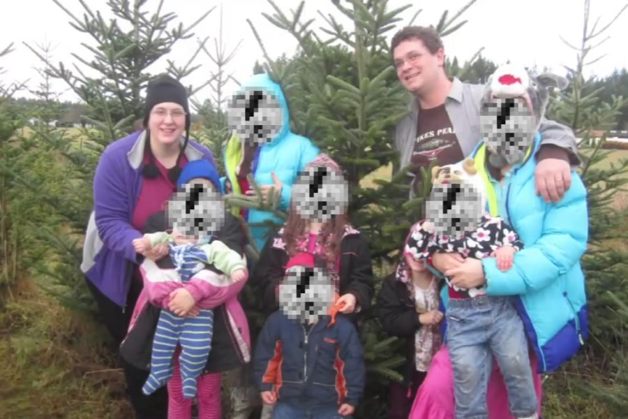 A large family poses together in front of Christmas trees at a tree farm. Most children’s faces are pixelated for privacy. Everyone is dressed warmly in winter clothing.