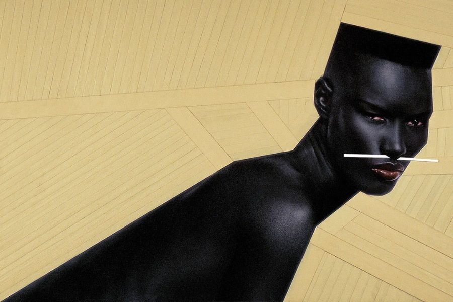 A model with geometric flat-top hair poses with high-contrast lighting, wearing a minimal black look and white sticks balanced across her face.