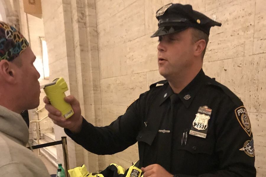 Police officer administering a breathalyzer test to a man indoors, holding a yellow handheld device.