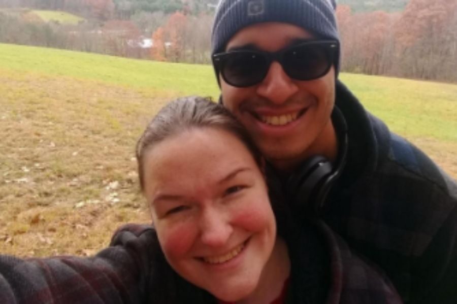 A couple smiles for a selfie in a field with rolling hills and autumn-colored trees in the background. The man wears sunglasses and a beanie.