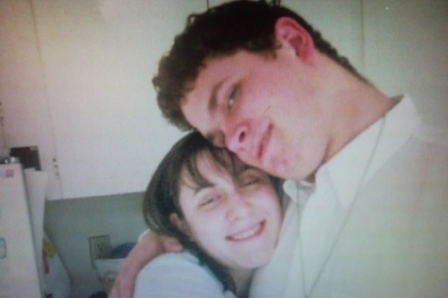A young couple embraces in a kitchen, both smiling at the camera. The image has a slightly grainy quality, suggesting it’s from an older photo.