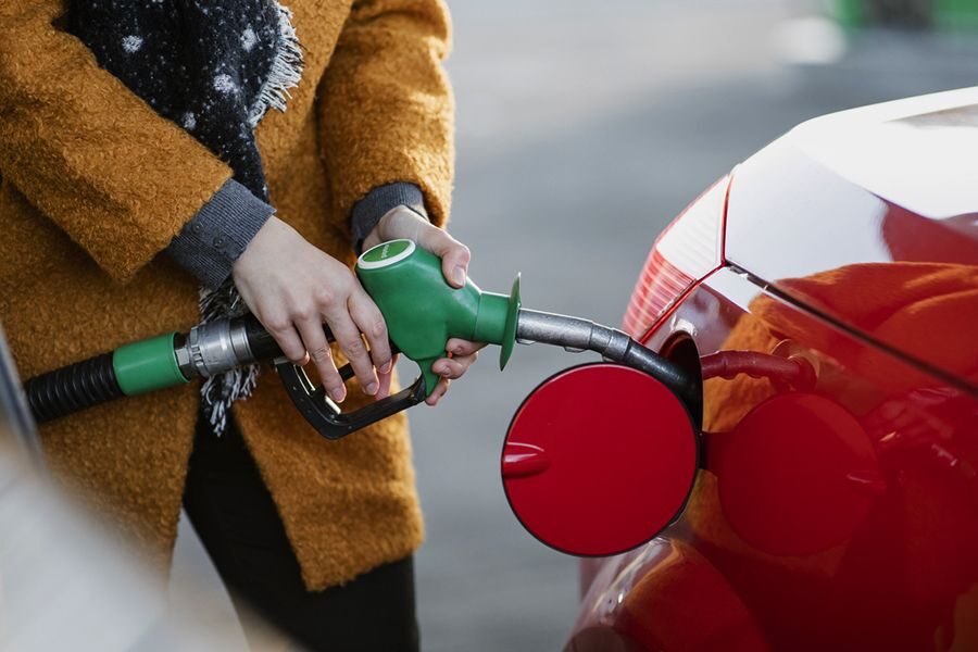 Person wearing a mustard coat fueling a red car with a green gas nozzle at a gas station.
