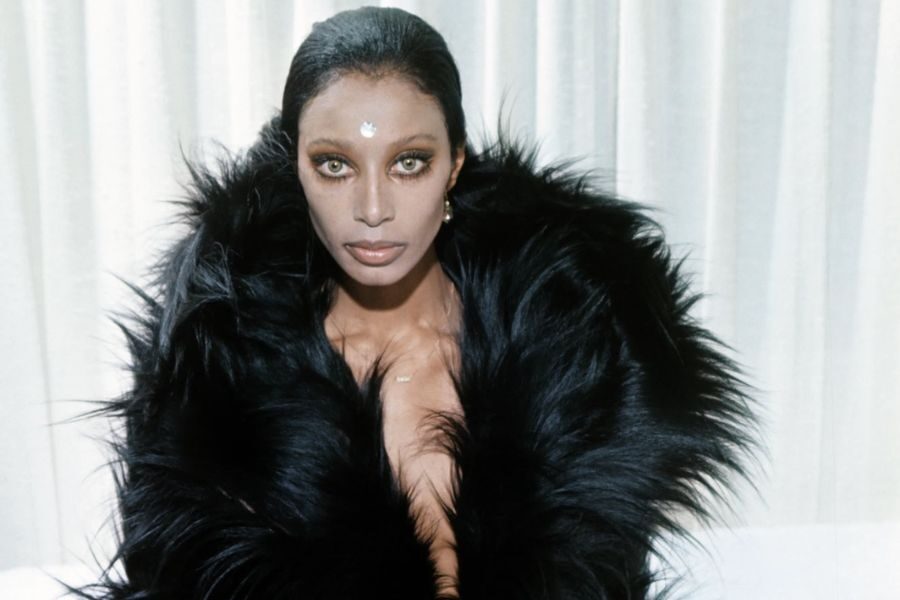 A model with striking eye makeup and a black fur coat poses indoors with a jeweled bindi and intense gaze.