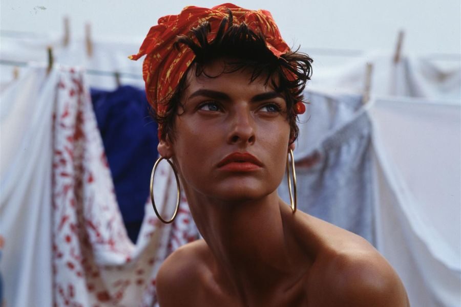 A model with short curly hair and red headwrap gazes off-camera while wearing large hoop earrings in front of laundry lines.
