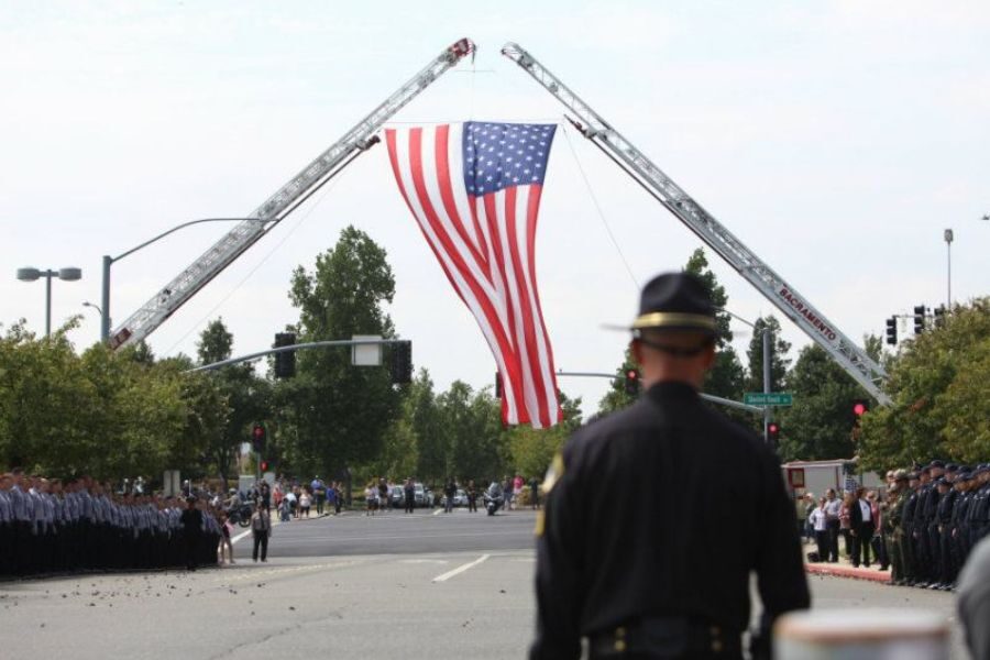American flag suspended over a city street by two extended fire truck ladders during a public event with officers in formation.