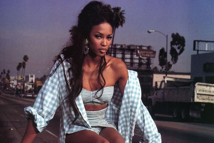 A model in a blue gingham matching set poses on an urban street, with voluminous hair and bold earrings.