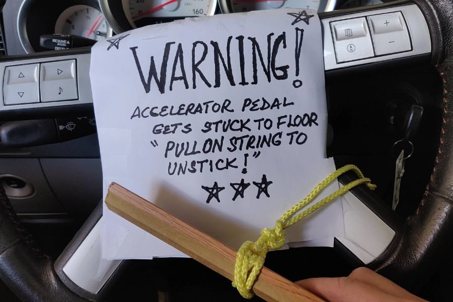 Handwritten warning sign taped to a car dashboard explaining the accelerator pedal sticks and must be unstuck using a string.