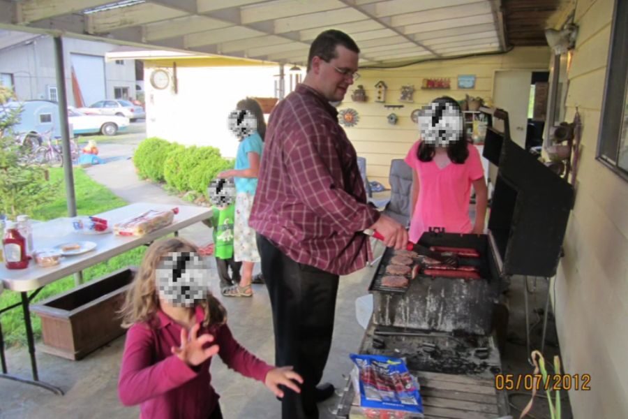 A man grills burgers and hot dogs on a black barbecue while surrounded by several children on a covered patio. The timestamp reads 05/07/2012, and the children’s faces are pixelated.