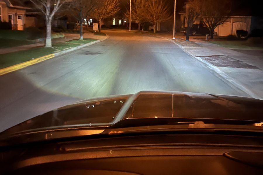 Car headlights illuminating a quiet residential street at night with trees and houses on both sides.