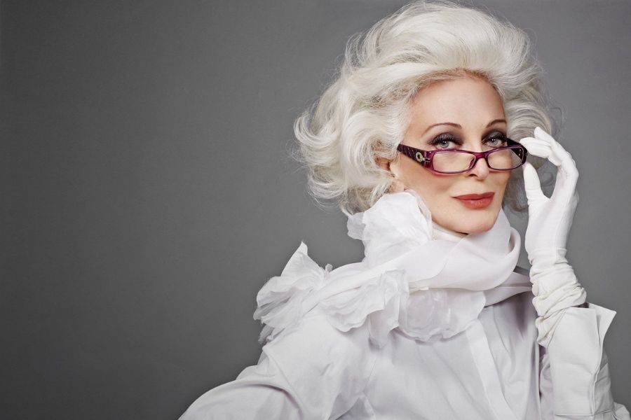 An elegant older woman with voluminous white hair wears ruffled white attire and purple glasses while striking a confident pose.