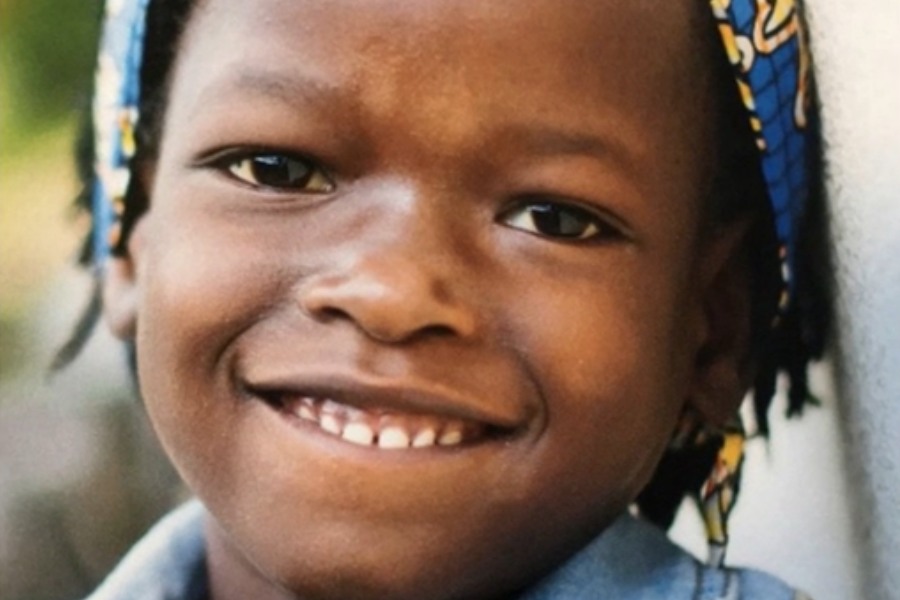 A close-up portrait of a smiling child wearing a colorful headband and denim shirt, with a natural blurred background.